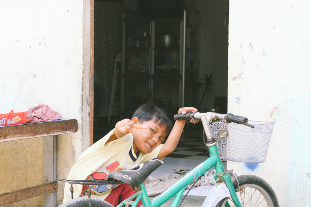 A little boy sits in front of his house holding a teal bike with a metal basket. He smiles and offers a peace sign to the camera man. 