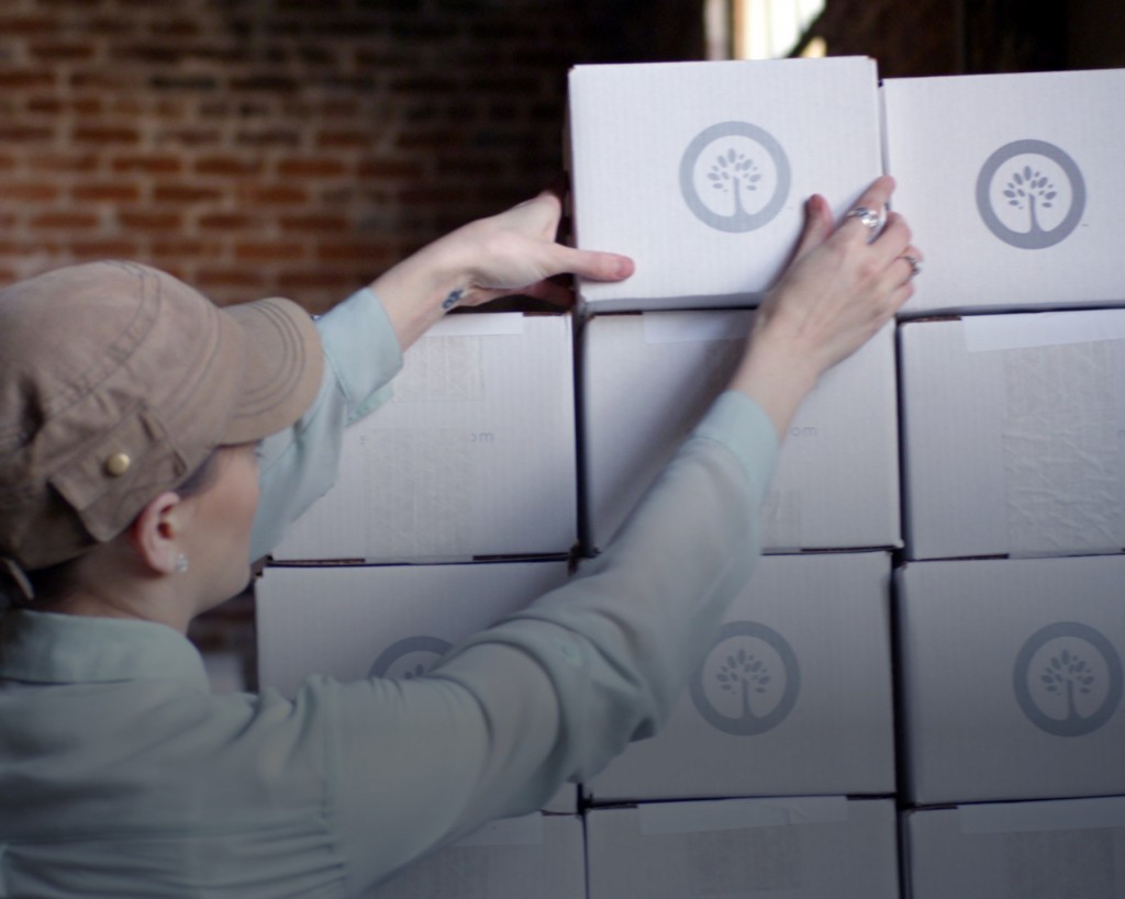 A female wearing a blue button down shirt and brown cap reaches for a white box with a gray Southtree logo. in the middle. The box is on top of a stack of similar boxes. In the background is a red brick wall. 