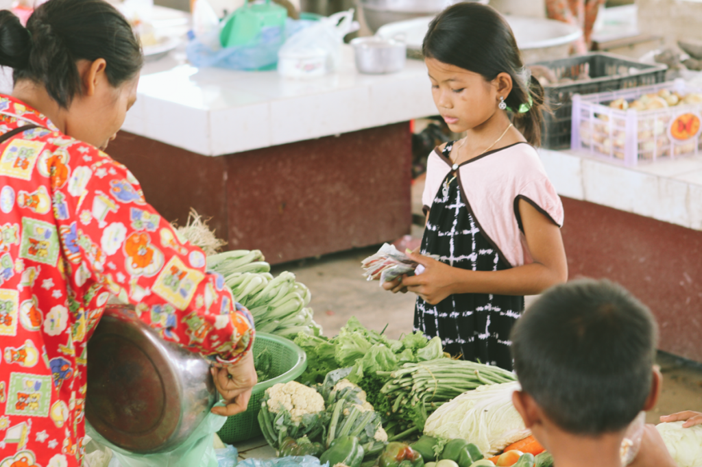 A little girl stands behind a table of fresh vegetables. She is counting a wad of cash, about to offer change to a woman purchasing fresh vegetables. Other vendors sit off in the background. 