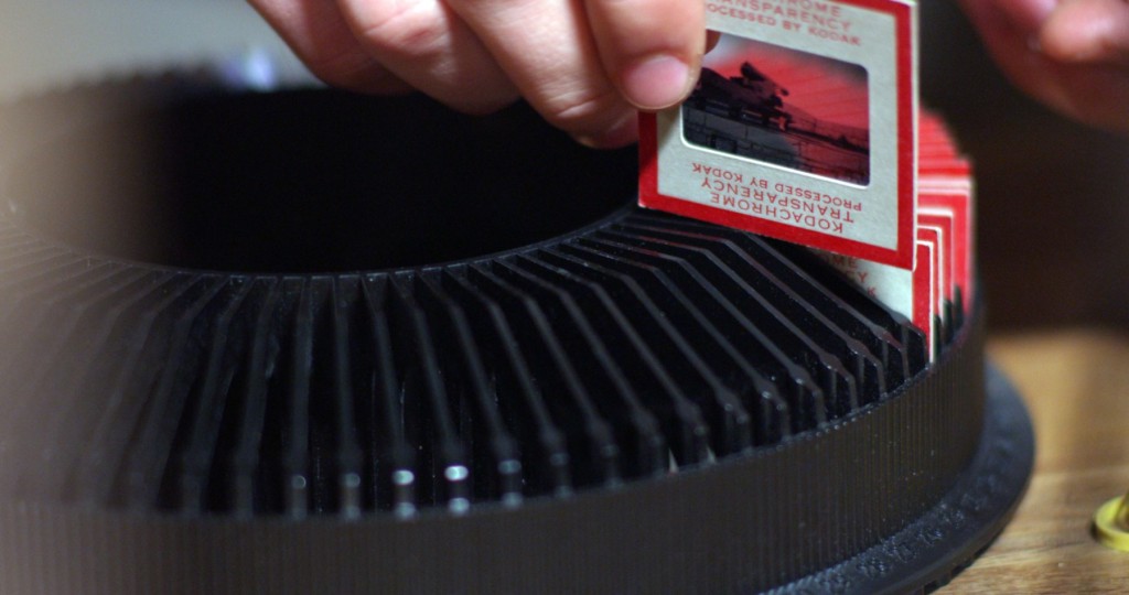 White slides with red pictures are being placed in a black slide carousal.  The image on the slide in the foreground is a of a bright print sunset over a lake. 