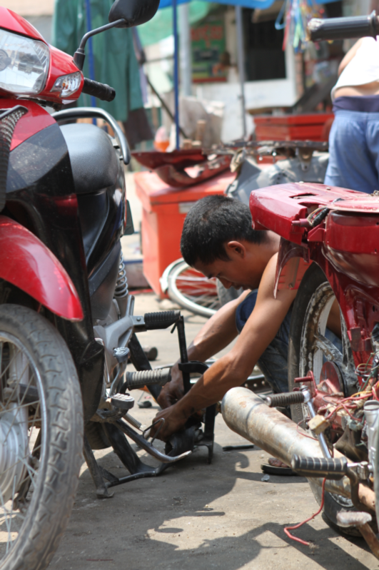 A young man works in the street to repair his motorcycle. The sun shines down on him. He is surrounded by other vehicles needing repair and stacks of scrap metal. 