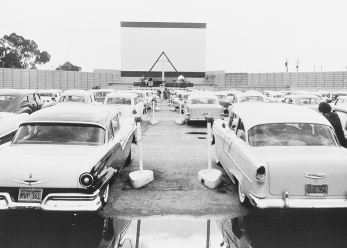 Black and white photo from the 50s of cars lined up at the drive in waiting for the film to start. 