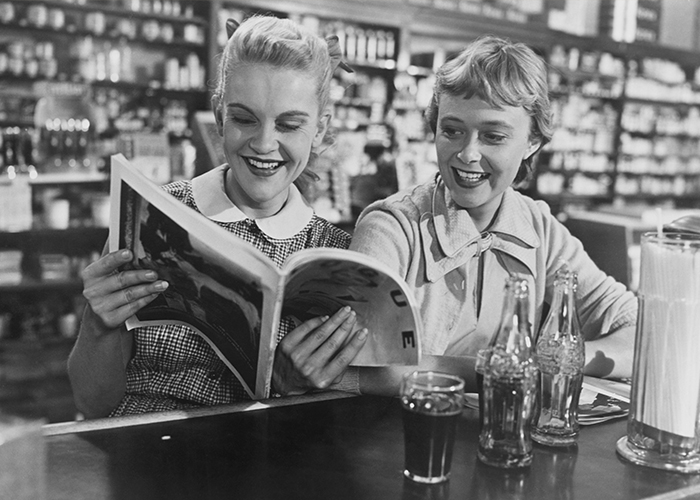 Black and white photo of two women in their 20s sitting at a soda fountain drinking Coka-Cola and reading Vogue magazine from the 1950's.