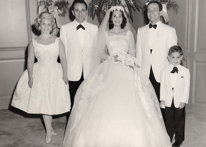 An iconic bridal party from the 60s. The bride stands in front in a princess chiffon  gown. A large large lace rose has been worked onto each of her hips.  To her right is her groom and two her left her father. Her mother stands next to her father in an icon 1960s dress -- fitted at the wait, flaring and ending right below the knee. Next to the groom is the ring bearer. All of the men are dressed in black slacks, white blazers, and black bow ties. 