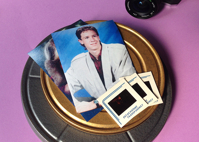 On a light purple background, sits a silver film reel case. Stacked on top is a gold film reel case. On top of that are 2 photos. The top one is of a young man wearing a cream jacket with large shoulder pads. It looks like one of the posed photos from the 80s. On top of that photo are three fanned out slides.