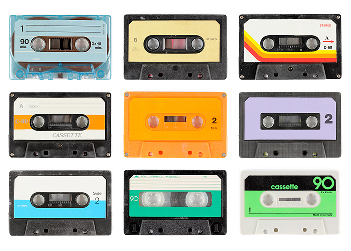 Nine different audio cassettes sit on a white seamless background. Their colors and designs vary from transparent aquamarine to opaque black with a cream label containing a yellow, orange and red stripe. 