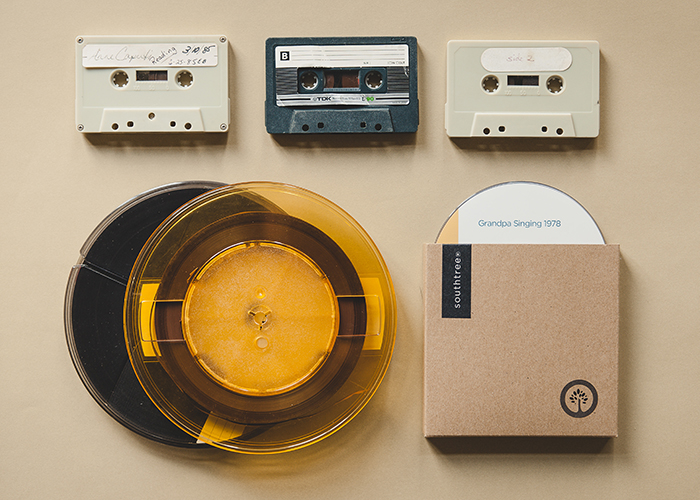 Various audio formats sit on a tan background. Three plastic audio cassettes, about the size of a deck of cards, sit in a line at the top. Under them sits an older audio reel and a CD titled, "Grandpa Singing 1978"