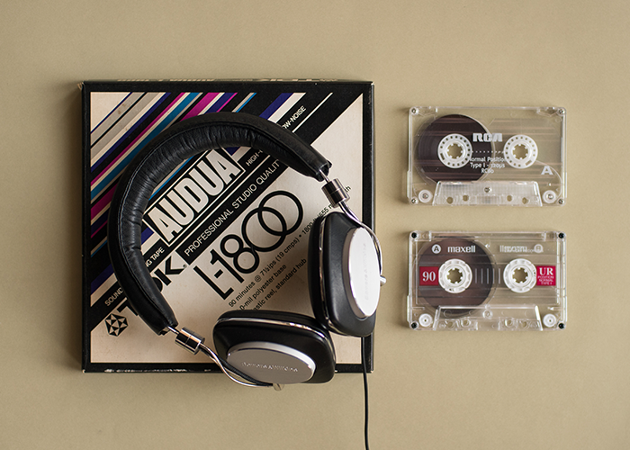 Black and silver headphones sit on an eggshell colored Audio box with magenta, eggplant, taupe and black stripes. To the right of the box sit two clear plastic audio cassettes. Everything is on a rich beige background.