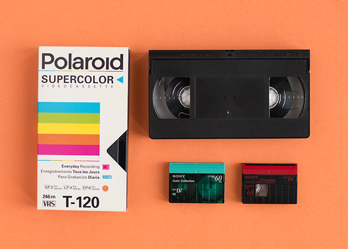 Four VHS tapes sit in a square grid on a seamless background the color of orange sherbet. On the left is a vertical standard VHS tape in a white case. The white case has a blue, green, yellow orange and pink stripe across the middle. At the top is says "Polaroid" in black san-serif text. The rest of the box is filled with black technical copy. To its right is another black plastic standard VHS but this one lays horizontally and is not in a case. Two horizontal black plastic mini 8mm VHS tapes sit beneath the un-cased VHS. One has a turquoise spine and the other a bright red spine. These items are shot from an arial point of view. 