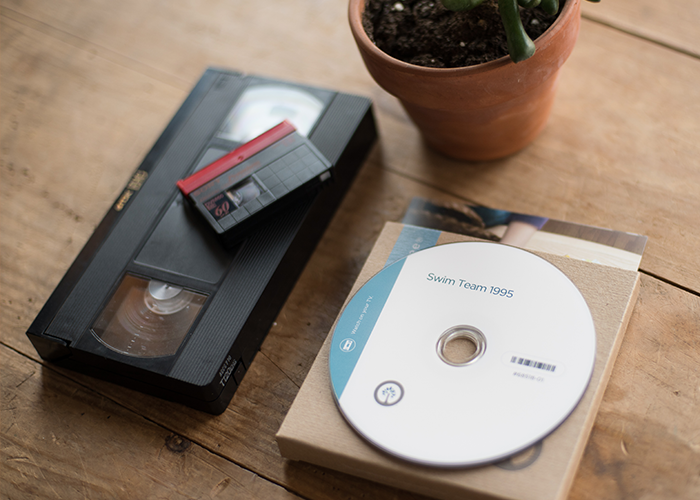 On the left side of a wood plank table, a standard black plastic VHS tape lays on its back. On top of it sits a black 8mm tape with a bright red spine.  Cut off at the top of the frame is a medium size succulent in a red terra-cotta pot. To the left sits a kraft paper southtree DVD case with a white CD on top. The CD says, "Swim Team 1995". This whole scene is shot from an arial vantage point. 