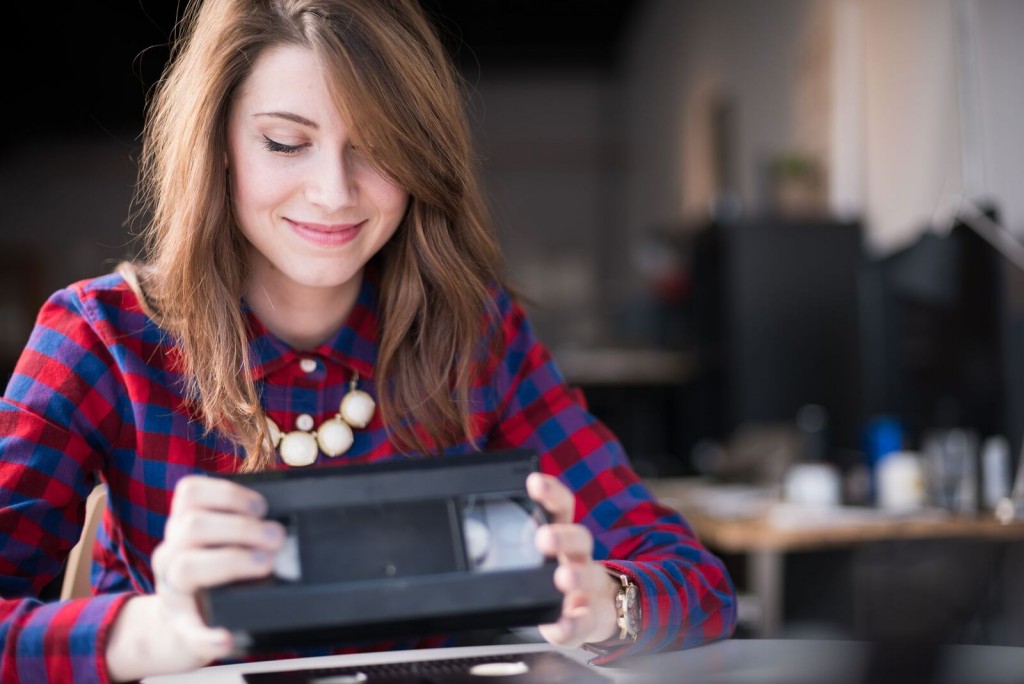 A young brunette female in a red and blue plaid shirt sits at her desk. She's smiling as she fondly looks down at the VHS she's holding in her hands.