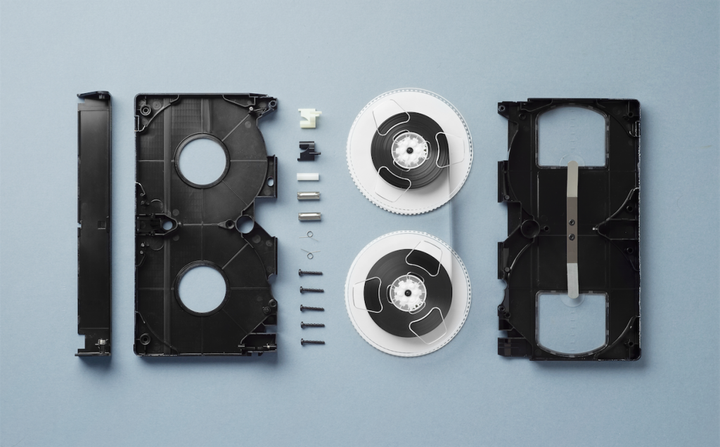 A disassembled VHS sits on a sky blue background.  The VHS's flap sits vertically on the far left of the shot, next to it is the plastic front of the VHS. Next to that sit a vertical row of the devices screws at bolts. Next to that are is the reels of film. The plastic back sits on the far right of the shot, bookending the composition. 