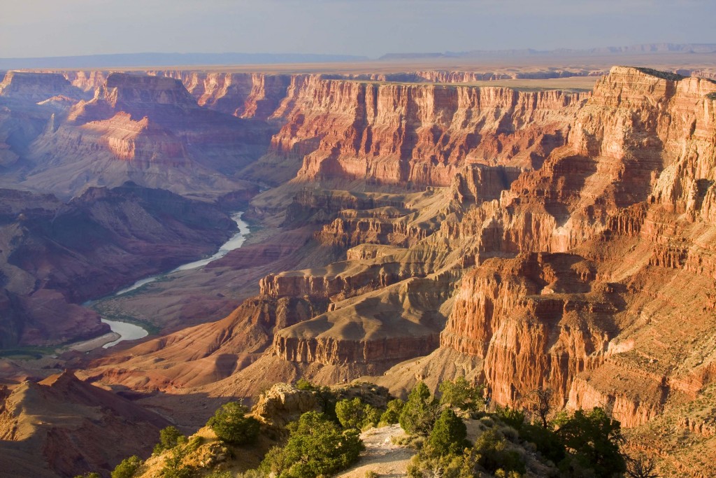 The Grand Canyon at dusk. Walls of pink stone line the large canyon. In the middle of the canyon flows a river. Green trees are sprinkled throughout. 