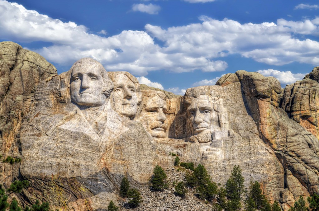 Mount Rushmore. 4 presidents faces - George Washington, Thomas Jefferson, Abraham Lincoln and Theodore Roosevelt - are carved into the side of a mountain in South Dakota. The base of the mountain is sprinkled with trees and a clear blue sky with scattered clouds lingers overhead. 